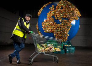Greg Wallace leaning on a trolley full of food in front of a structure of a planet made of food, part of a food waste campaign from WRAP's Food Waste Action Week